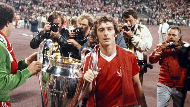 Trevor Francis holds the European Cup after winning it for the first time with Nottingham Forest in 1989. The trophy is on the left of him. Francis wears a red Forest shirt with a red scarf around his neck. He is surrounded by photographers and fans can be seen celebrating in the crowds in the distance