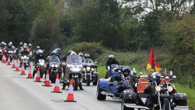 A convoy of motrcycles, with traffic cones sectioning off one lane of the road. There are three-wheeled and two-wheeled vehicles, some of which are carrynig flags.
