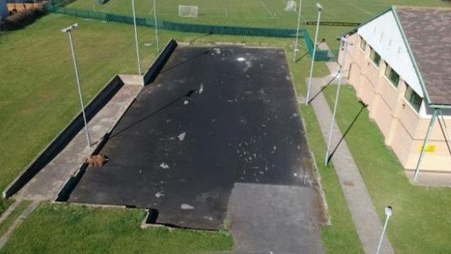 An outdoor asphalt pitch in poor repair with floodlights, surrounded on all sides by grass and to the right a brick building.