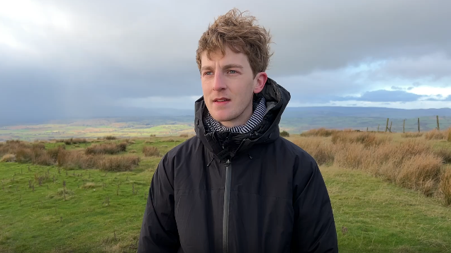 A man stands on grass on top of a hill, he is wearing a black hooded jacket. He has long light brown hair that is being blown around by wind.