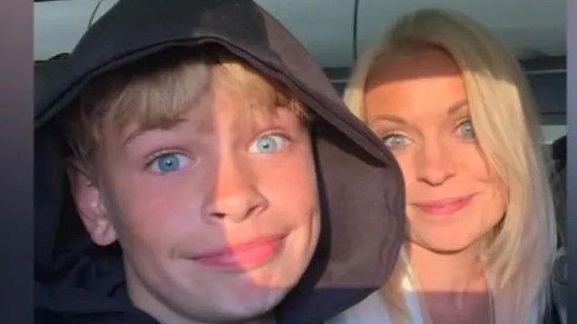 Mikey Roynon with his mother, Hayley Ryall. Mikey is on the left of the frame. He is blonde, has blue eyes and is wearing a black hoodie. He is smiling. Hayley is sat next to him smiling - she also has blonde hair and blue eyes. 