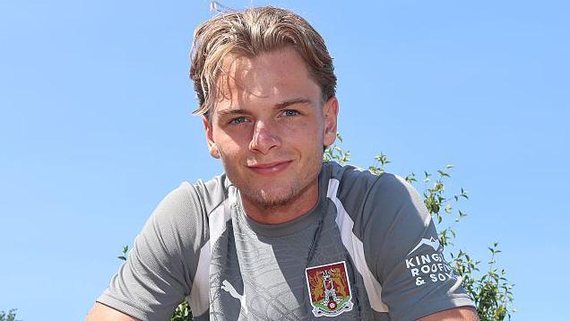 A smiling Logan Briggs wearing a grey Northampton Town training top faces the camera