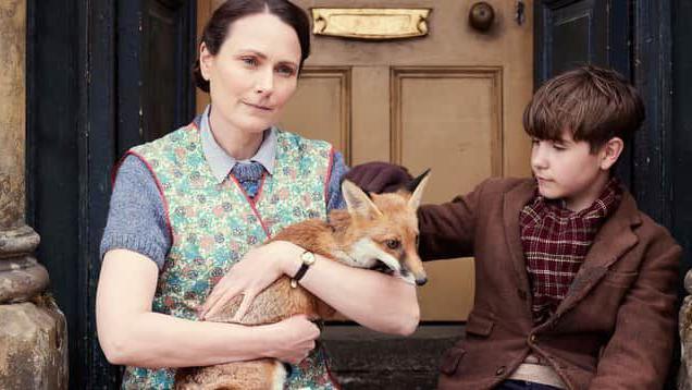 A boy sits in front of a door, next to a woman holding a fox cub in her arms. His left arm rests on the animal's head. The woman has her brown hair tied up and looks into the distance.