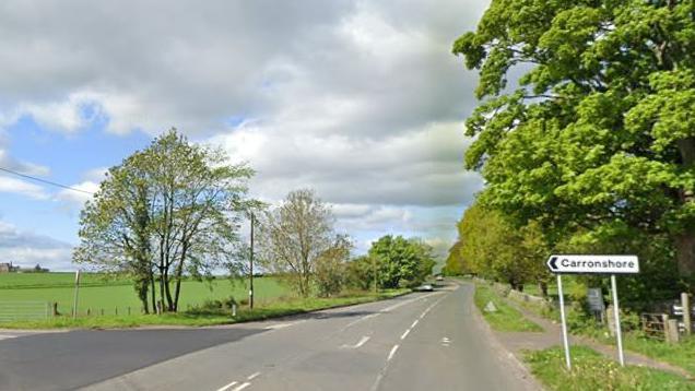 A stretch of road surrounded by fields with a sign that says Carronshore facing a junction .