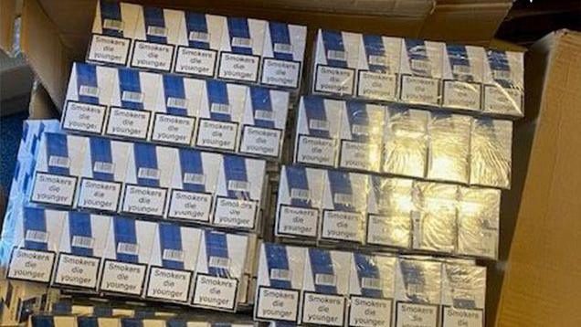 A cardboard box containing dozens of blue-and-white boxes of counterfeit cigarettes, bearing the warning 'Smokers die younger', and wrapped in clear plastic.