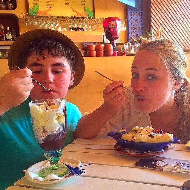 A young Scott, in a green t-shirt and a sun hat, tucks into a massive knickerbocker glory ice cream as he sits in a cafe with his sister Katie, who is enjoying a banana boat. A bar area is behind them with shelves of glasses and drinks and a price list on the wall.