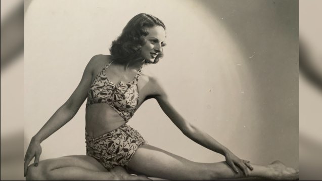 Carol in a dance cropped top and shorts in a split dancing pose. She has a side parting and curls with lengthened arms away from her body in a dance pose.