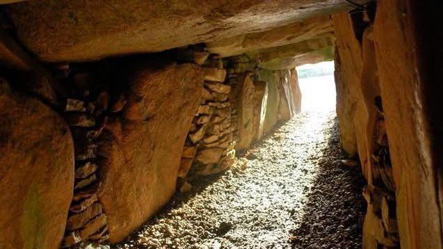 The entrance to La Hougue Bie. It is taken from the inside of a cave. The rocks are brown. There is sunlight coming in.