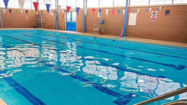 Water glistens on an empty indoor swimming pool. Lane are marked out and red, white an blue bunting hangs above the water.