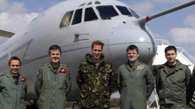 Five men are posing in front of a large grey aircraft, on the left is Grant Lawrie, he is smiling in his RAF olive green uniform. In the middle of the group is a young Prince William, he is wearing a camouflage outfit.