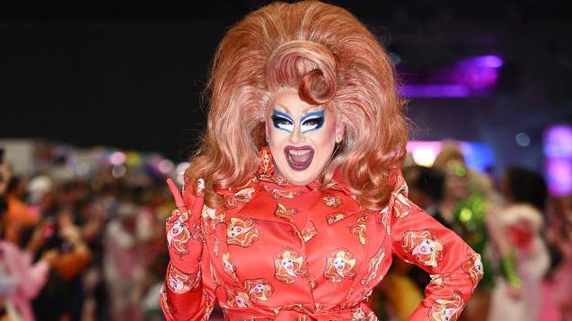 Drag artist Porkchop with a red outfit on and wearing a large red wig with blonde streaks.