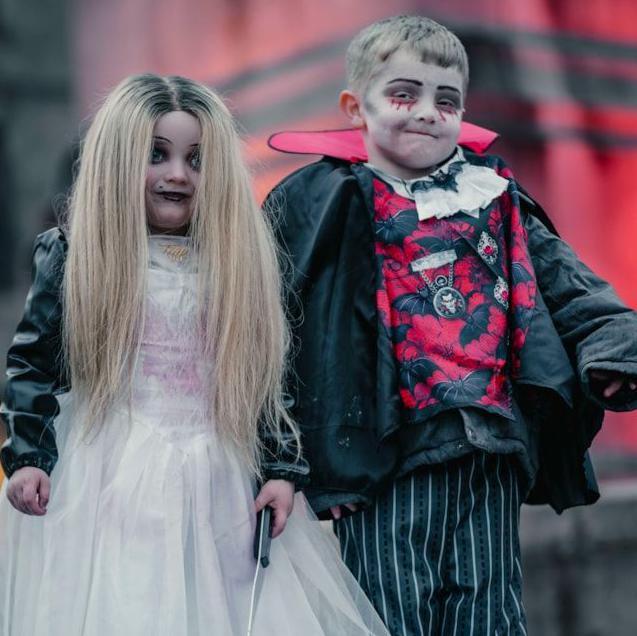 A young boy and girl in Halloween costume - the girl has long blonde hair, white face paint with dark circles around her eyes and wears a white dress. The boy is dressed as a vampire, mostly in black with a red collar and a red jumper depicting black bats.