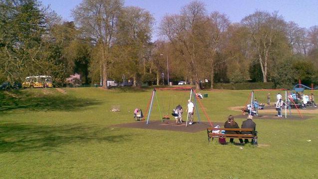 Large grass park with trees in the background. There is a playground in the foreground with swings and a toy train. There are children playing on the equipment and some adults are sitting on a bench. There is a yellow and white ice cream van in the background.