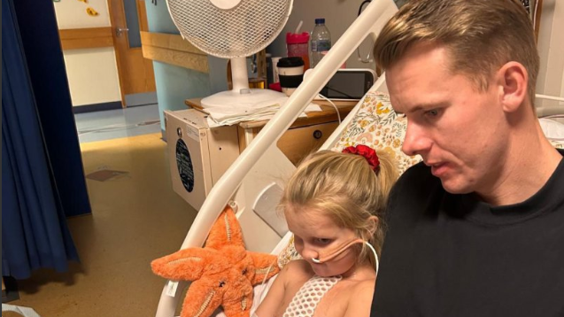 Tom is sitting in the hospital bed with Graycen as he reads her a book. He is wearing a black top. Gracyen has hospital tubes in her nose and a post surgery plaster on her chest. There's a cuddly star fish next to her.