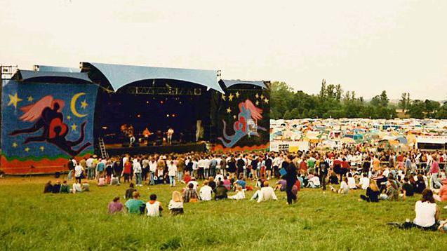 A main stage at the festival, with a band performing. There is a roof with a semicircle edge over the stage. There are people sitting on the grass watching the stage. A campsite is visible to the right.