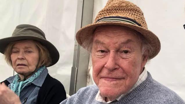 A close-up selfie-style photo of two people seated side by side in an outdoor event space. In the centre, Timothy West is sitting slightly behind the camera holder, wearing a woven brown hat and a light grey sweater over a collared shirt. On the left, Prunella Scales is partially in frame, seated further back. She is wearing a darker hat, a scarf, and a jacket, and is positioned slightly turned away from the camera.