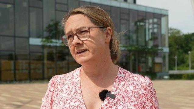 Helen Harrison with long brown hair tied back wearing glasses looking to the right of the camera. She is wearing a red and white dress to which a microphone is attached. There is a glass-fronted building, the Corby Cube, behind her.