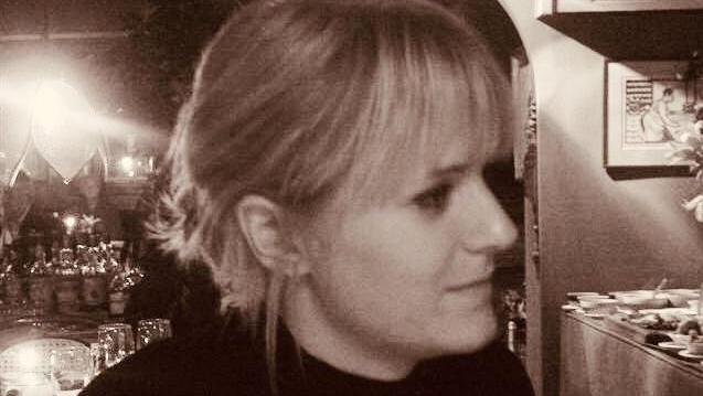 A black and white image of a side profile of a woman with tied back blonde hair wearing a black jumper. She is inside a room which has lights, a picture and flowers in.