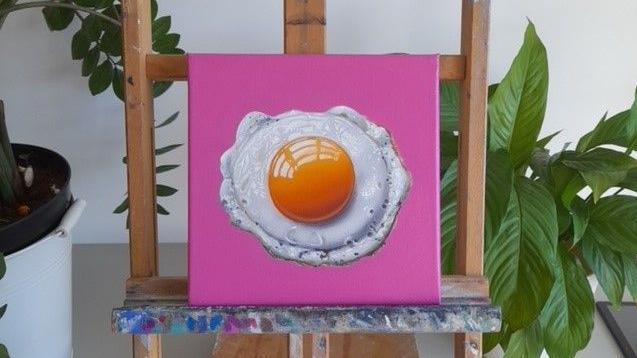 A sunny-side up on a pink canvas, popped on a small easel with paint marks on the wood. Two plants can be seen in the background.