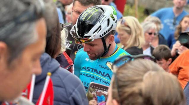 Mark Cavendish wearing a pale blue cycling top and white helmet in a crow of fans signing autographs.