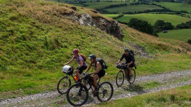 Three cyclists are riding uphill on a small stone path. Surrounding them are rolling green hills that stretch into the horizon. 