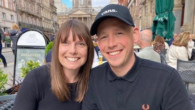 Hannah has shoulder length brown hair and is wearing a black top. Shaun has a black baseball cap and a black Fred Perry shirt. He is clean shaven and has a shaved head.