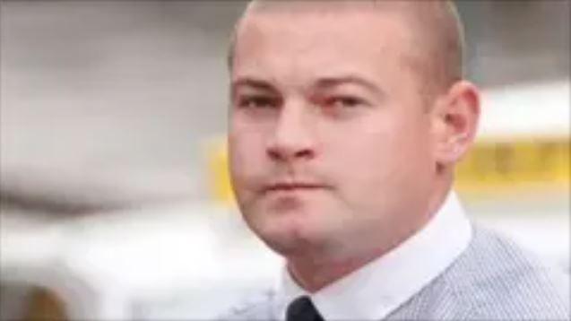 A man with a shaved head wearing a shirt and dark tie looks at the camera. He is standing outside but the background is blurred. 