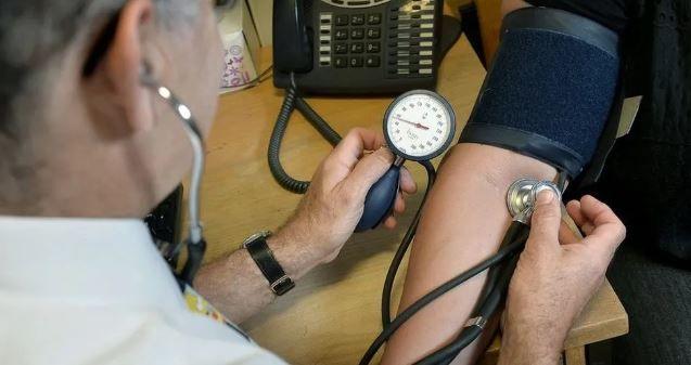 A doctor sitting at a desk taking a patient's blood pressure. He is holing a stethoscope against the patient's are, which has a dark blue pad wrapped around the elbow area secured with Velcro. The doctor is holding a pressure gauge in his other hand.