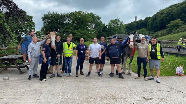 A group of volunteers, some in high visibility vests, pose with two horses in a picnic area.