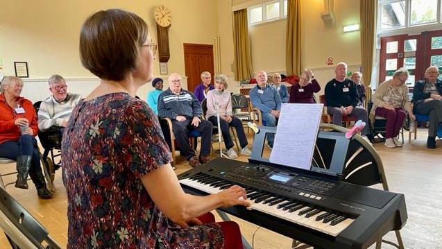 A women playing an electronic keyboard with people sat on chairs in a semi circle around her in a village hall.