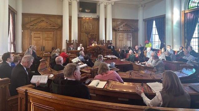 Councillors sitting a wood-panelled room. In the background is the top table with five people sitting in ornate chairs. In front of them, in a U-shape, there are lots of people sitting in pews with notebooks in and paperwork in front of them. The large windows are letting a lot of light in.