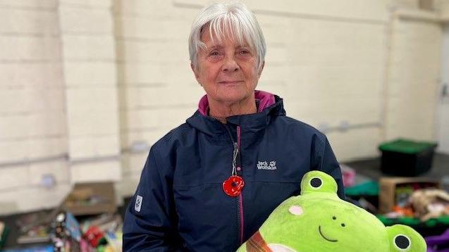 Female charity worker stands with a fluffy green coloured toy. She's got short grey hair and wears a navy jacket.