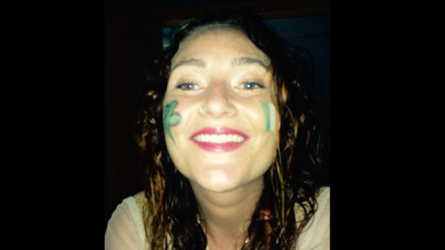 A woman smiles into the camera. She has face paint on her cheeks of a shamrock and a tricolour flag. 