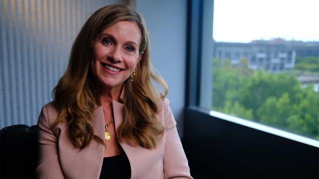 Julie Inman Grant in a pale pink jacket and black blouse smiles as she looks at the camera. She is sitting next to a window.