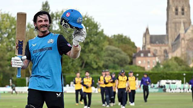 Worcestershire's Rob Jones salutes the crowd with his helmet and bat as he walks off the field having scored a hundred in a 50-over match. He is wearing the club's light blue and black One-Day Cup kit with the opposition players and Worcester Cathedral in the background.