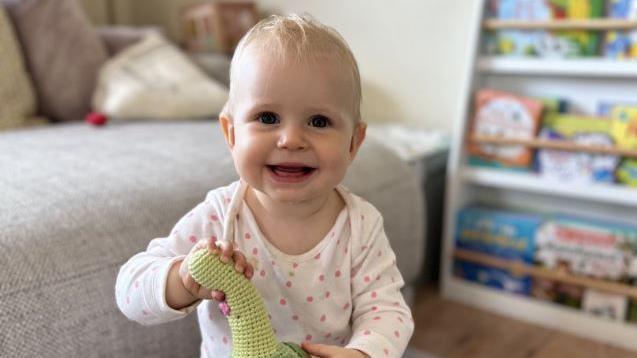 Olivia stood in her front room holding a soft toy. She is looking directly into the camera. Behind her is a sofa and a children's book shelf filled with books.