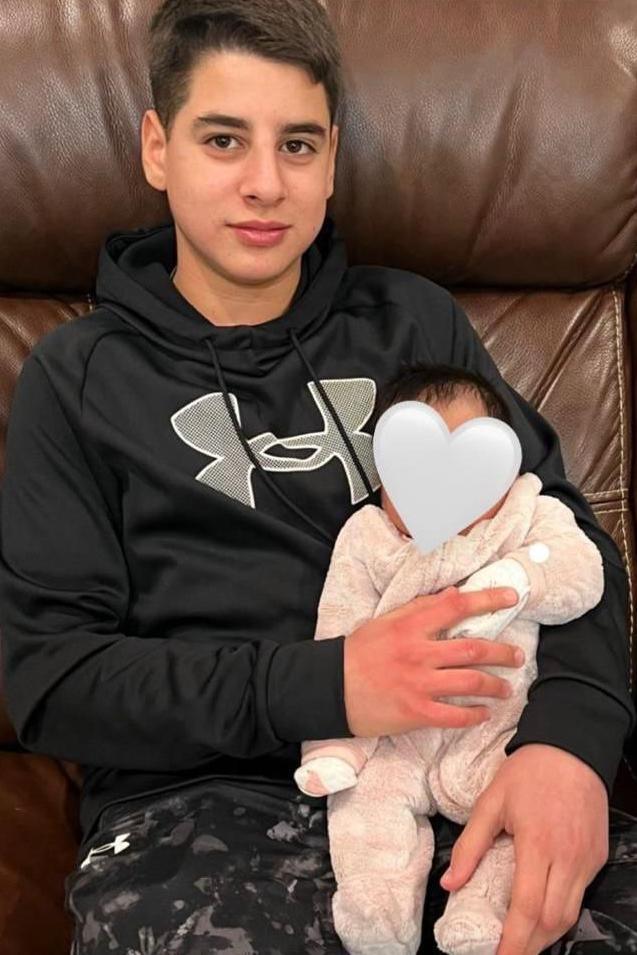 Mohammed is sitting on a leather sofa and holding a small baby whose face has been covered with a heart emoji. He is smiling and wears a black hooded jumper and patterned trousers, and the baby is in a pink fleece onesie. 