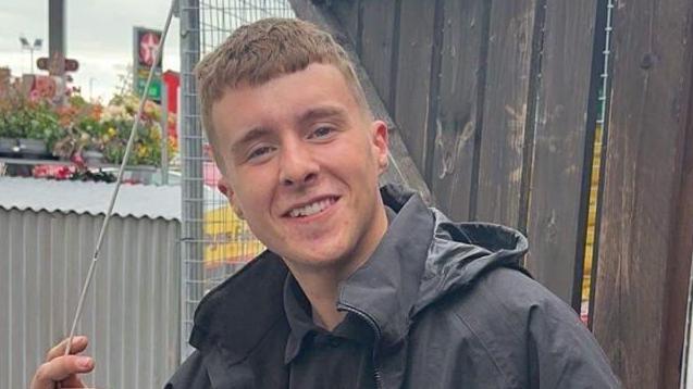 Matthew McGrath has short brown hair, he is wearing a black polo shirt and black raincoat. Behind him is a brown wooden fence, a corrugated iron structure and a petrol station forecourt.