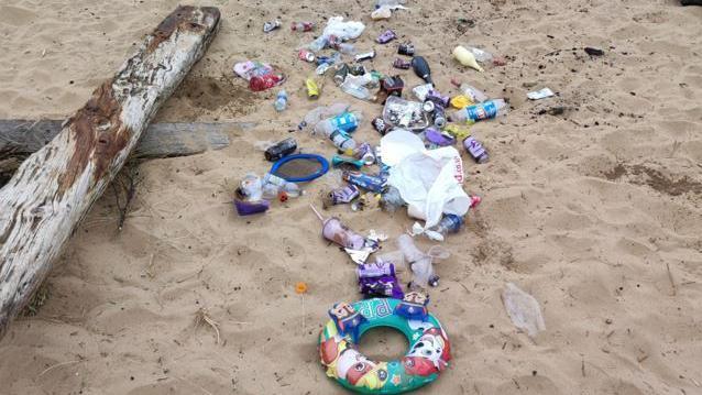 Child's rubber beach ring, plastic bottles, paper strewn on a beach beside a log of wood. 