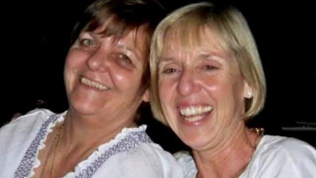 A lady with short dark hair leans her head towards a lady with blonde short hair. Both are looking straight into the camera with wide smiles. Both are wearing white tops. 