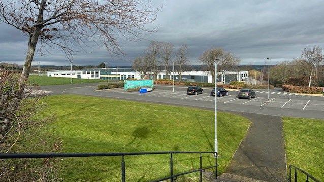 Prudhoe High School viewed from the top of steps into the school groups is a one storey building in white and grey cladding set over a large area. In front is a carpark around the school are playing fields 