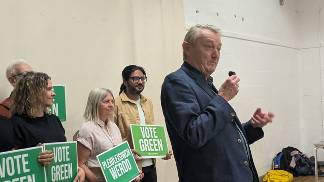 Anthony Slaughter in a blue suit holding a microphone with four people behind him holding vote Green signs in English and Welsh.