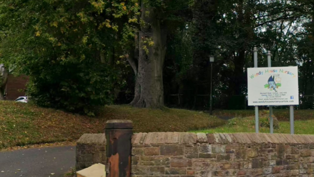 A Google Streetview screenshot of the entrance to Wendy House Nursery in Carlisle. A colourful sign with a drawing of a house is erected behind a stone wall, next to a drive, surrounded by grass and trees, with fallen leaves around them. In the distance is a red-brick building.