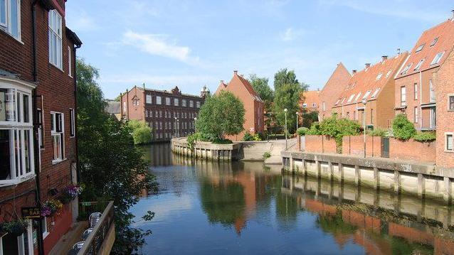 A river weaves past homes and a pub in the middle of Norwich on a sunny day.