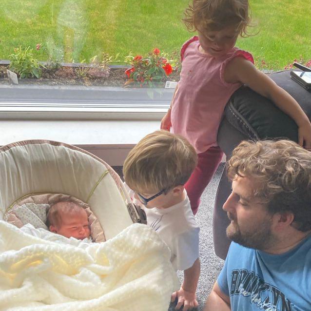 Baby SS lies in a moses basket next to a window with a view to a garden. Her dad Tommy, wearing a blue t-shirt, and brother and sister are next to her. 