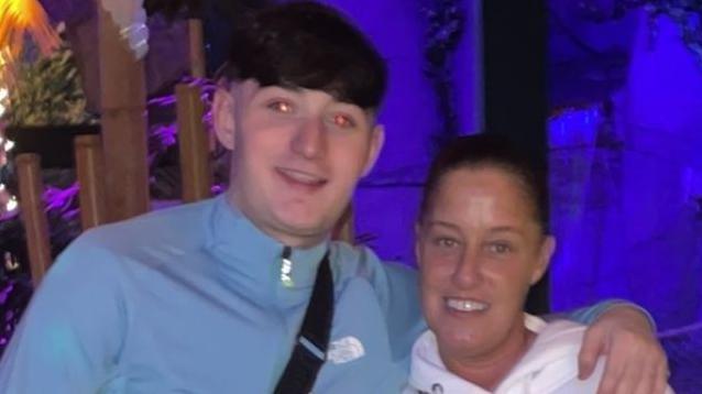 A man with short dark hair smiles at the camera. He wears a light blue tracksuit top and a cross body bag and has his arm around his mum, who is shorter than him. She has her dark hair tied back and wears a light coloured hoodie and smiles. 