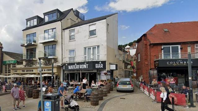 A series of cafes on Sandside in Scarborough with outdoor seating areas and people walking in the foreground on a paved area.
