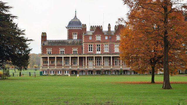 A red-brick manor house with a small tower and columns sits behind a wide area of grass with trees on either side. 