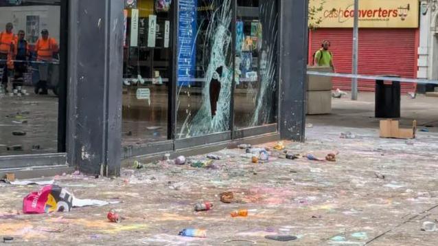 Rubbish, filth and lush bath products of various colours strewn across the ground in front of a shop with a broken window. Police tape surrounds the scene. 