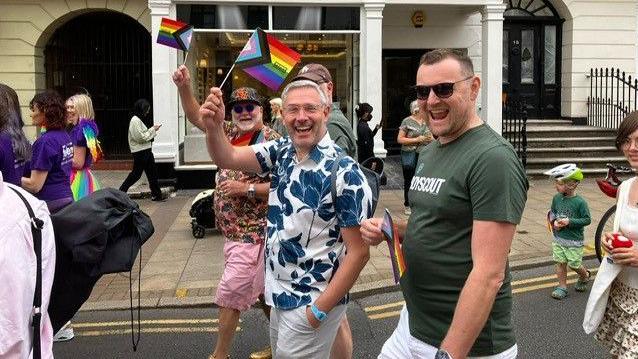 Three men wave at the camera also waving small flags as they walk in the parade. They have short sleeved shirts on with one in a T'shirts. and they are wearing shorts.